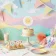 A toddler's birthday table set against a colorful paper backdrop, with matching invitations, a round cake topper and bunting flags all sharing the same bright flat-illustration style and p