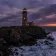 A lighthouse on a rocky cliff at bruised purple dusk, a single warm amber window glowing in the tower, a low sun smoldering on the horizon, waves breaking on dark rocks below.